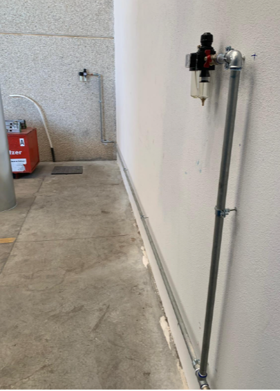 Tubi metallici e regolatori del filtro dell'aria montati su una parete interna bianca in un ambiente di officina.