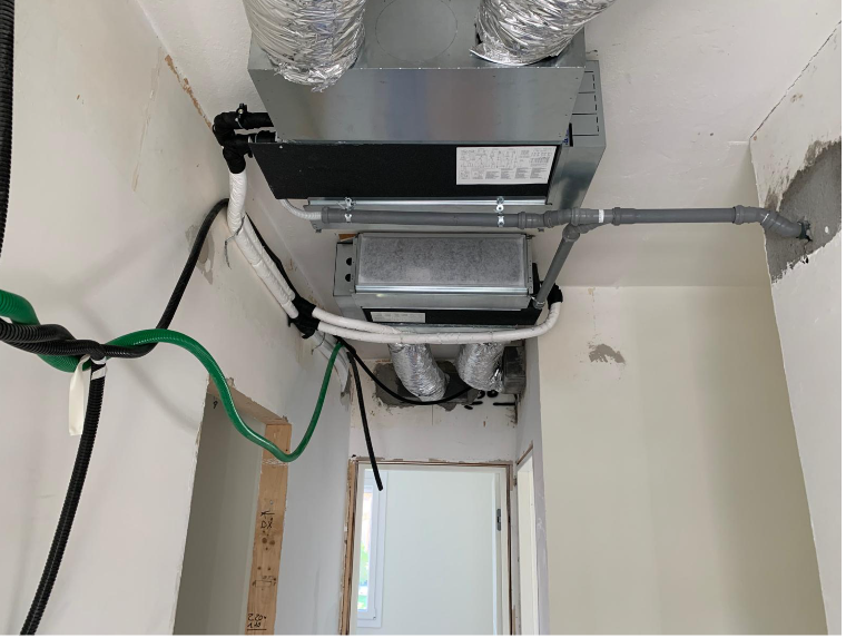 Unità di climatizzazione a soffitto con condotti metallici, tubi grigi e cablaggio elettrico in un corridoio interno non rifinito.