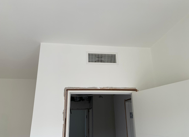 Una griglia di ventilazione rettangolare montata su una parete bianca sopra una porta con cornice non rifinita.