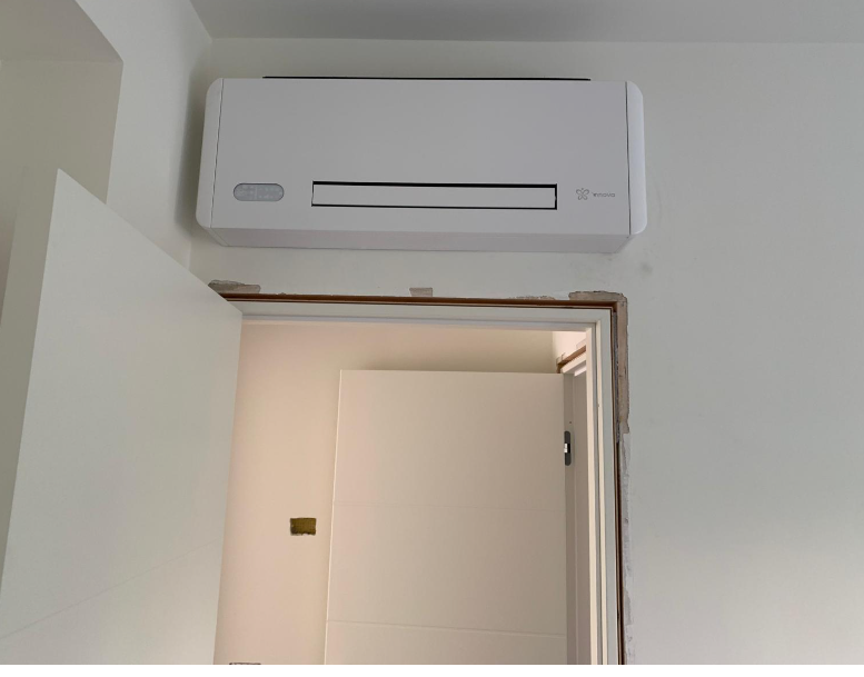 Un condizionatore d'aria bianco a parete installato sopra una porta in una stanza con pareti e porta bianche.