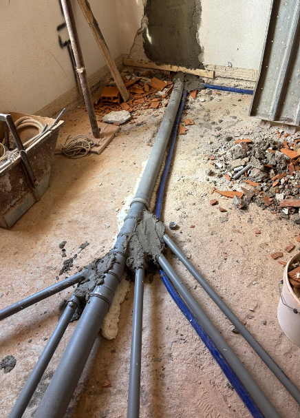 Il pavimento di un cantiere edile mostra tubi di drenaggio in PVC grigio, collegati tramite raccordi e rinforzati con malta grigia.