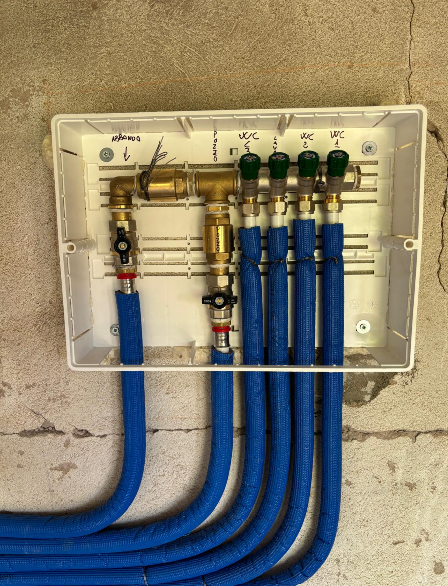 Un collettore idraulico con diversi tubi isolati di blu collegati a valvole in ottone all'interno di una scatola di derivazione a muro bianca.
