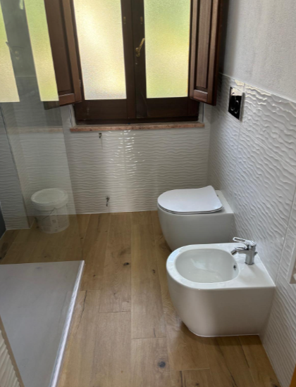 Un bagno moderno con WC e bidet sospesi di colore bianco, pavimento in legno e doccia con box in vetro vicino a una finestra.