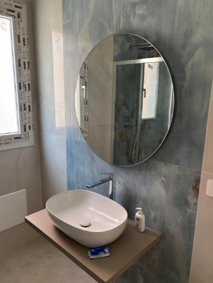 Un mobile da bagno con lavabo ovale bianco su mensola marrone, rubinetto a parete e specchio rotondo.