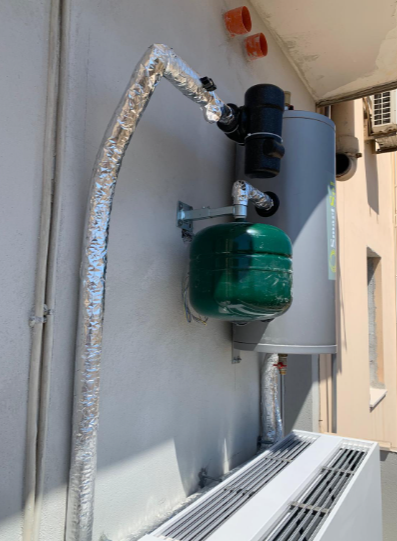 Un cilindro verticale grigio con un vaso di espansione verde annesso, montato su una parete esterna sopra un'unità di condizionamento dell'aria bianca.