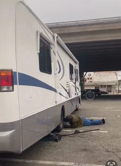 Man working on RV tire, under a bridge. Grey pavement, tow truck in background.