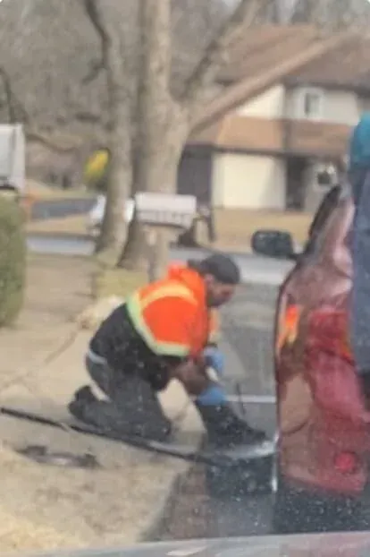 Person kneeling, wearing orange safety vest, working on a vehicle near a residential street.
