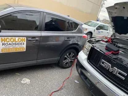 A grey car jump-starting a white GMC truck. Red jumper cables connected. H Colon Roadside sticker on the car.
