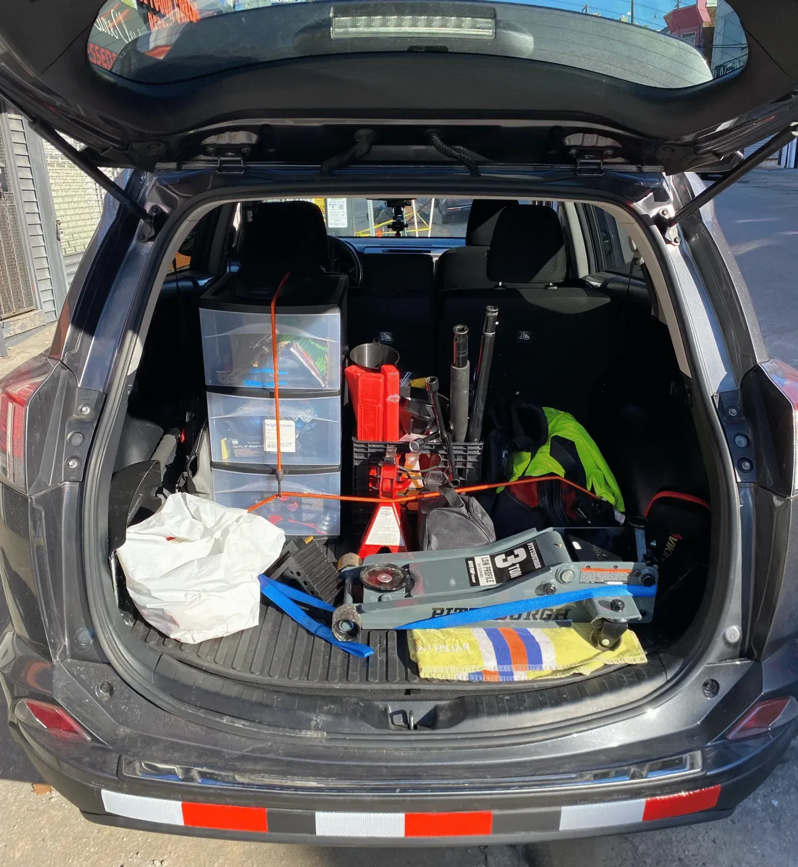 Back of a gray SUV filled with equipment: tools, plastic bins, a red machine, and other gear.