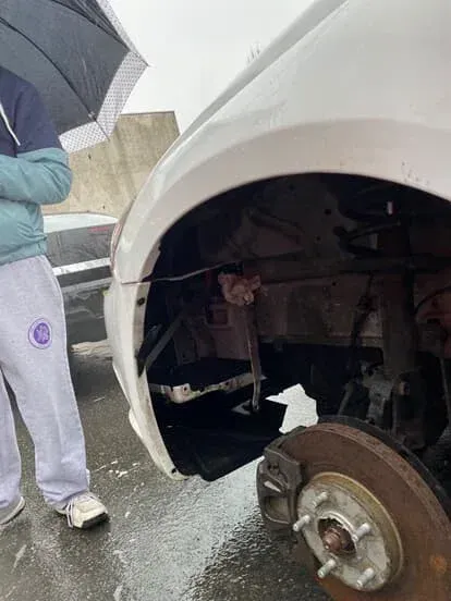 White car with a missing front wheel, person holding umbrella nearby.