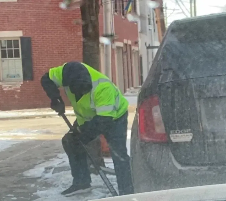 Person shoveling snow next to a dark car, wearing a neon green jacket. City street.