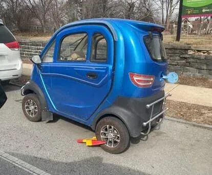 Blue, micro-sized car parked on a street with a wheel clamp; rear-mounted charging port.