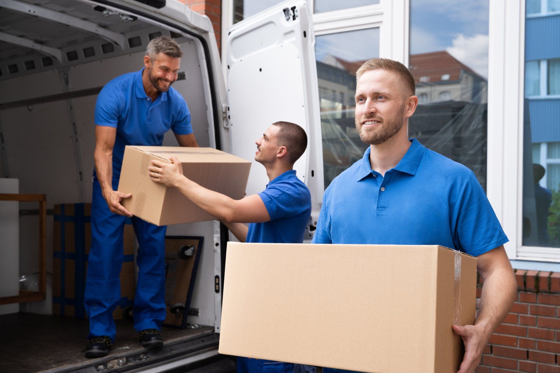 Tres mudadores vestidos de azul descargan cajas de una camioneta blanca, sonriendo al aire libre.