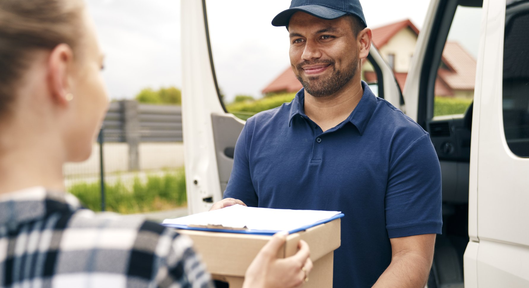 Un repartidor entrega un paquete y un portapapeles a una mujer, afuera de una camioneta blanca, sonriendo.