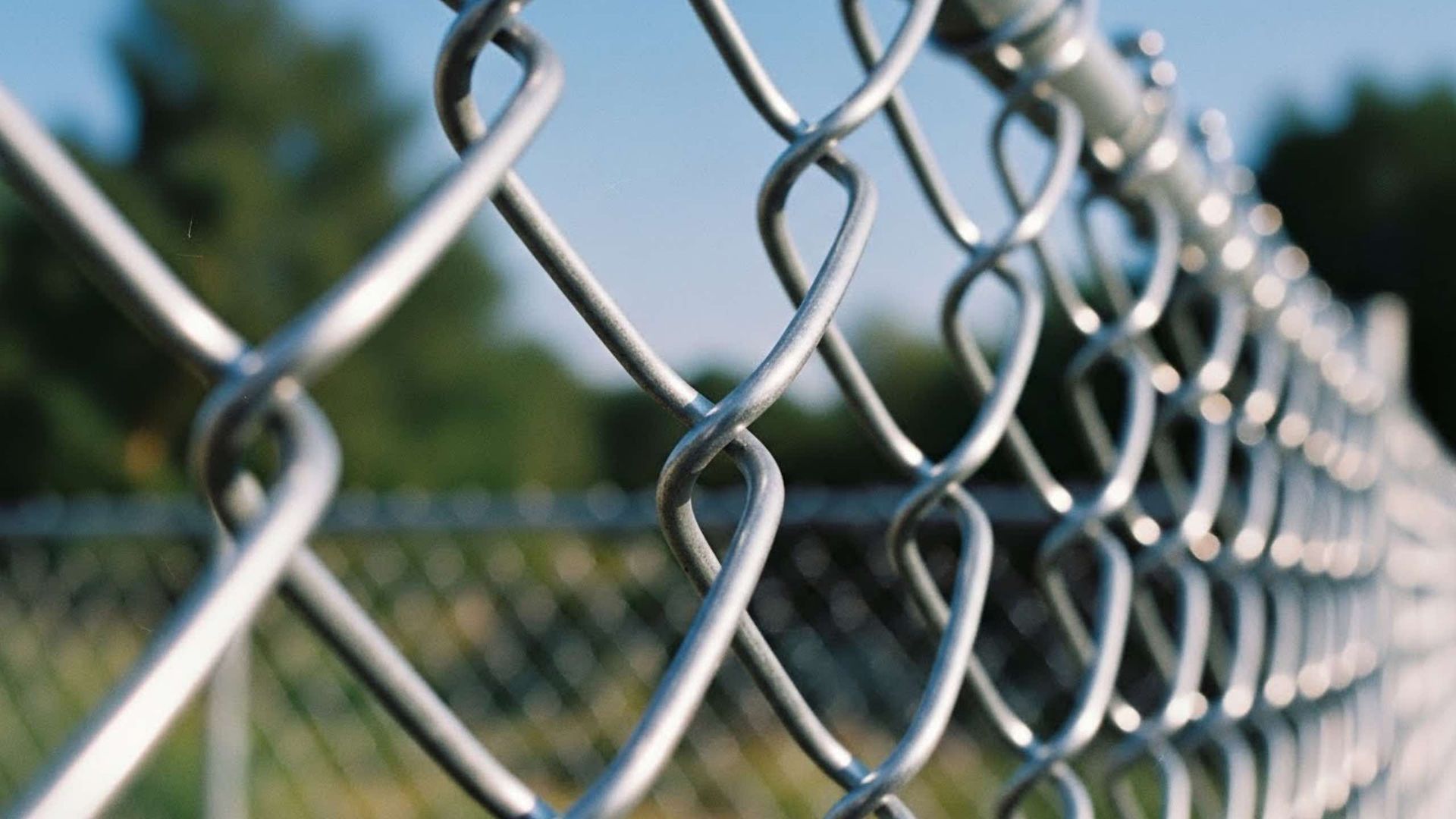 A close-up, angled view of a galvanized steel chain-link fence against a soft-focus background of trees and blue sky.