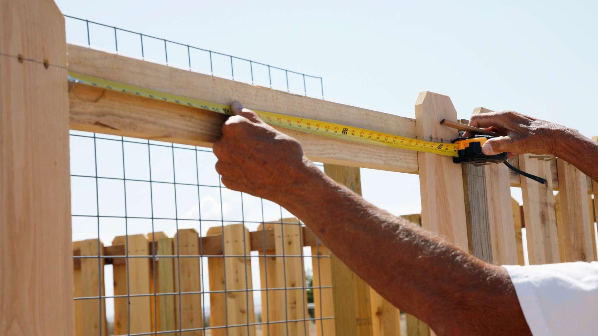 Person measuring a wooden fence with a yellow tape measure outdoors on a sunny day.