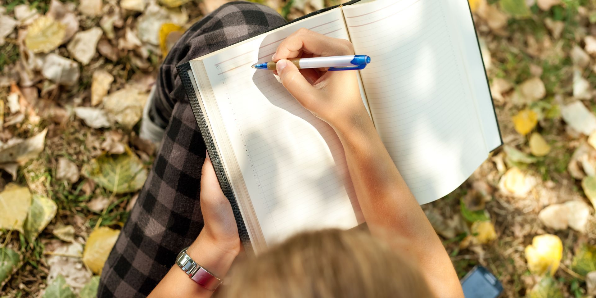 Person writing in a notebook with a blue pen while sitting on the ground covered with yellow leaves.