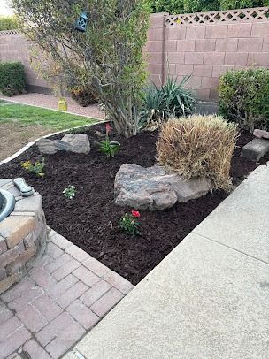 Backyard garden with rocks, mulch, and brick patio edging, by a brick wall and lawn.
