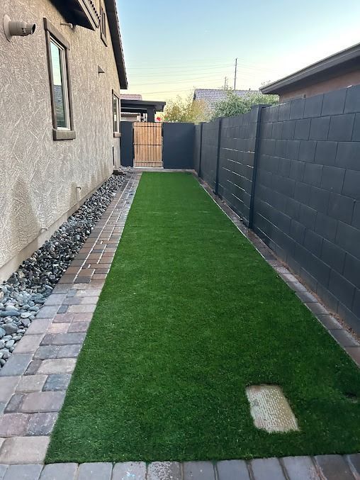 Narrow backyard with black fence, green turf, and brick pathway.