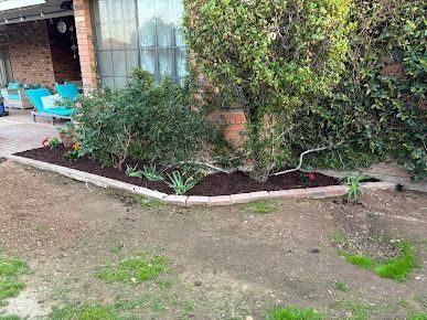 Brick-edged garden bed with dark mulch, green plants, and brick wall, in front of a home.