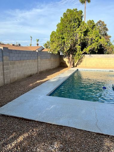 Backyard pool with concrete edging, surrounded by gravel and a block wall, under a blue sky.