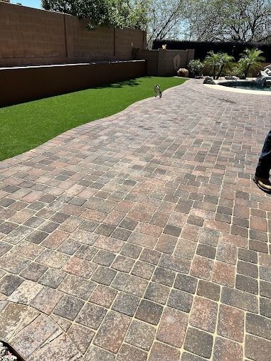 Brick patio next to green artificial grass in a backyard, brown wall in background, sunny day.