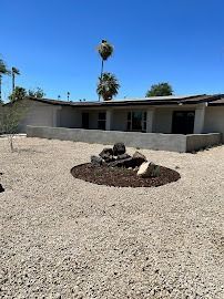 A low, single-story house with a rock and wood fire pit in a gravel yard on a sunny day.