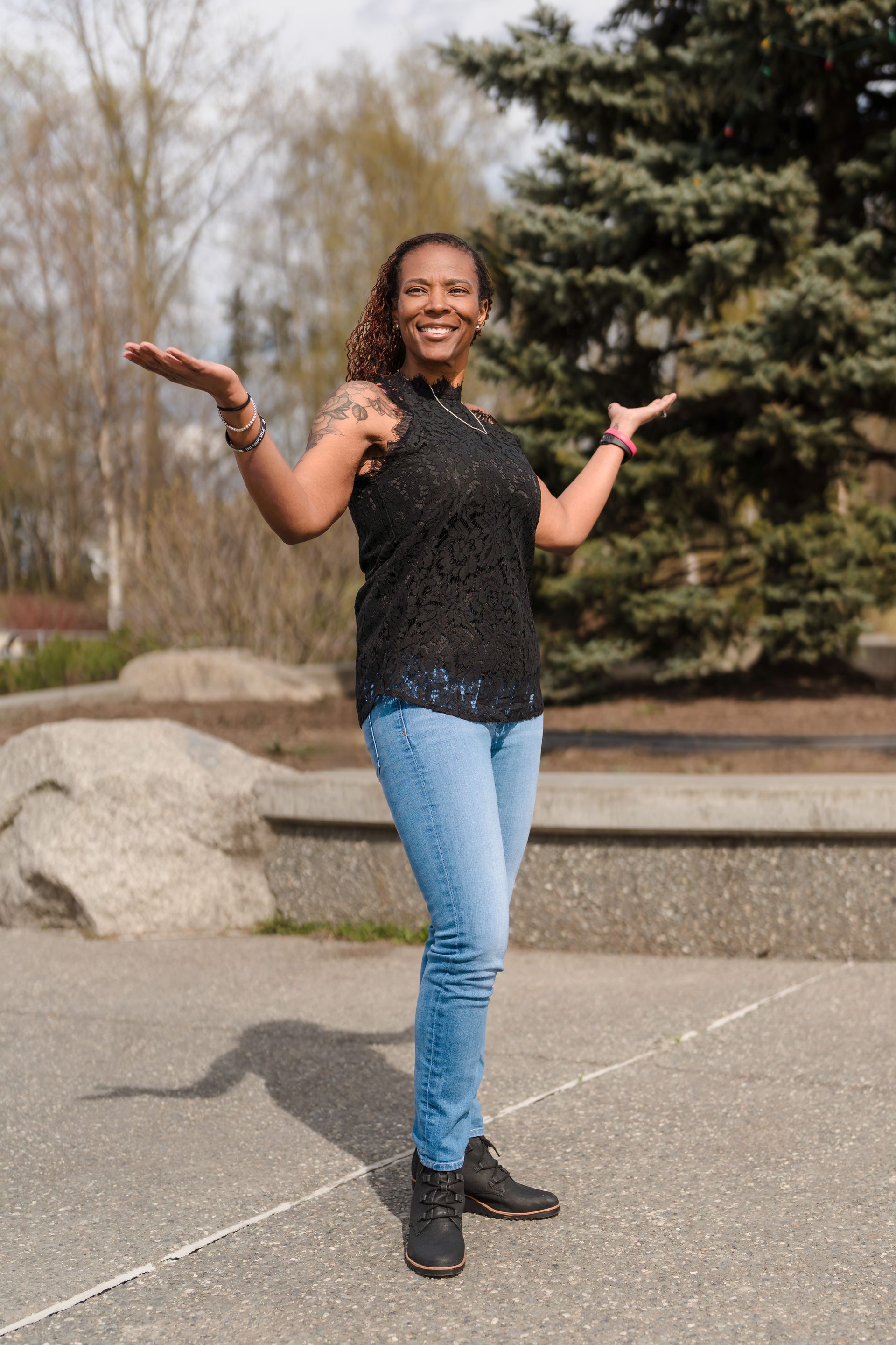 Woman with open arms, smiling, wearing jeans and black top, standing outside.