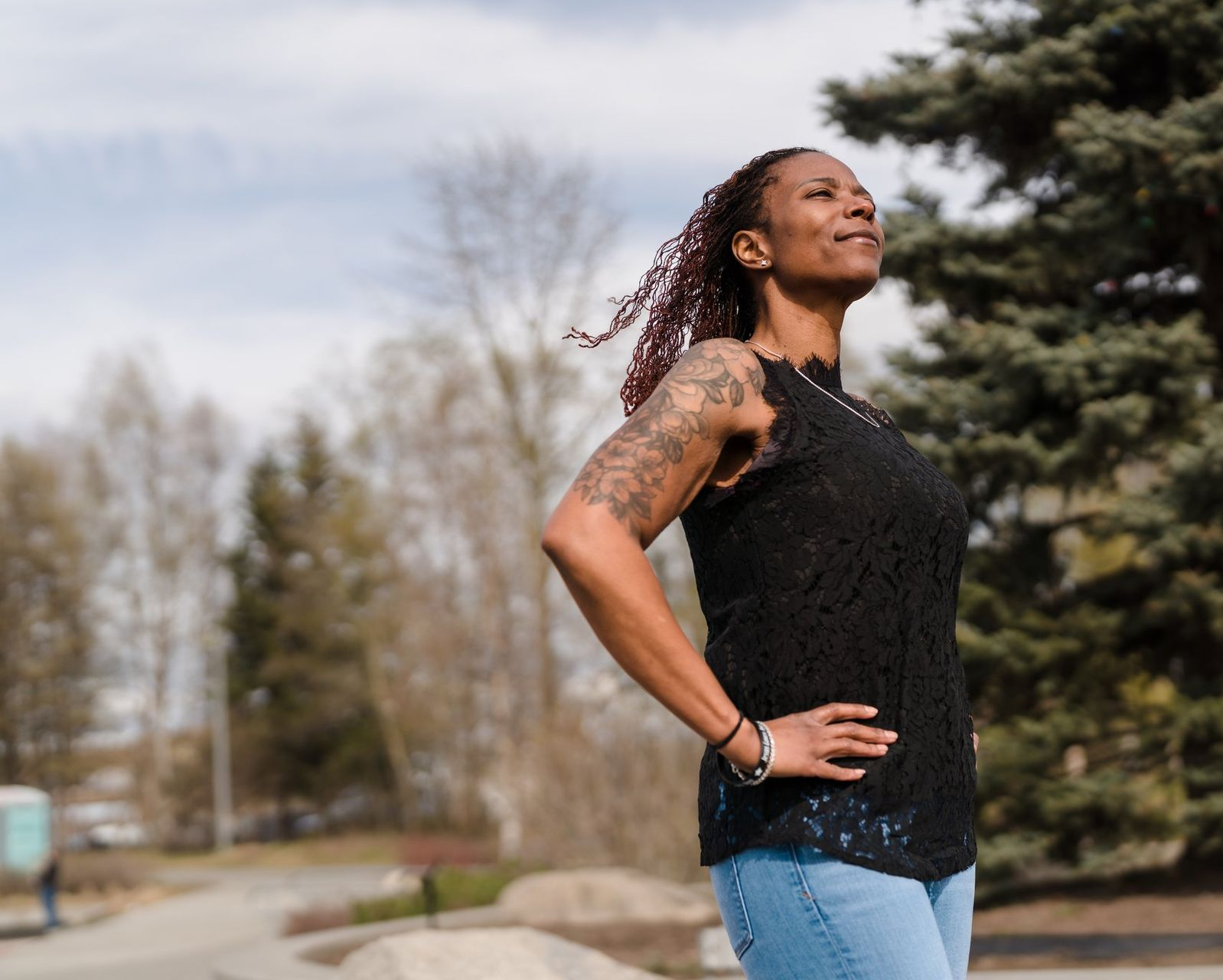 Woman in black lace top, arms on hips, looking up, outdoors.
