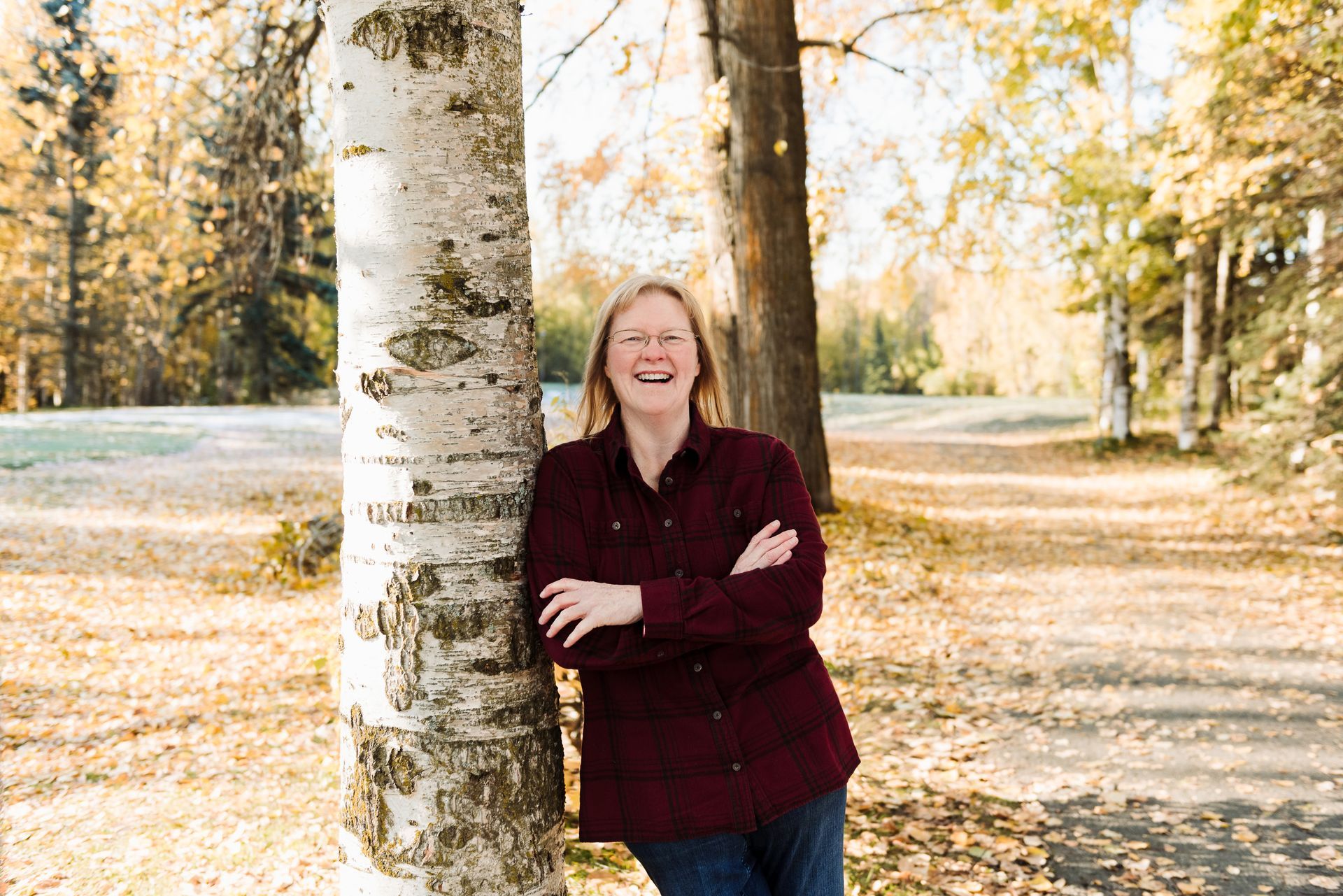 Woman in a burgundy shirt leans against a birch tree in a park, smiling. Autumn leaves are scattered on the ground.