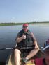 Man in a canoe gives a thumbs up on a sunny day. He wears a life vest and a red hat, paddling in a calm lake.