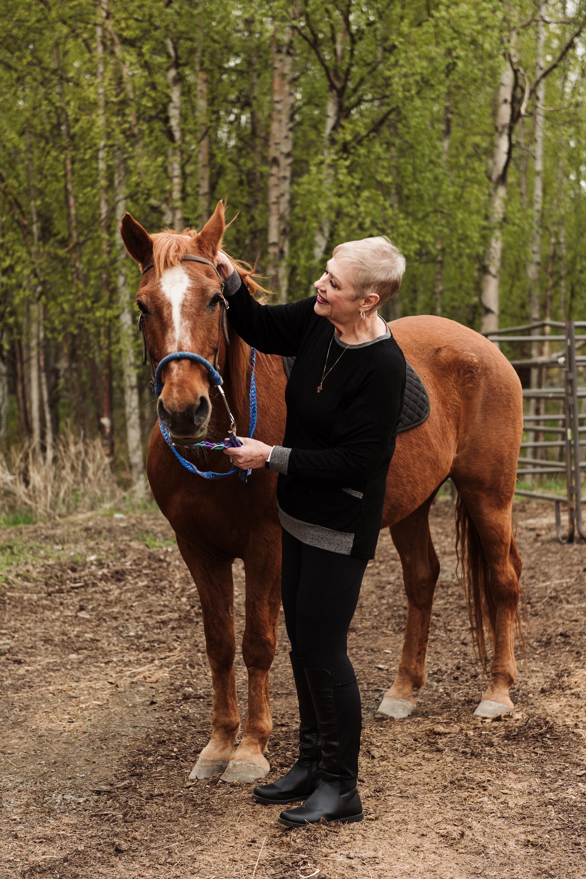 Woman petting a brown horse outdoors. Trees in background, holding reins.