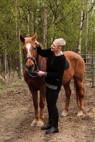 Woman petting a brown horse outdoors. Trees in background, holding reins.