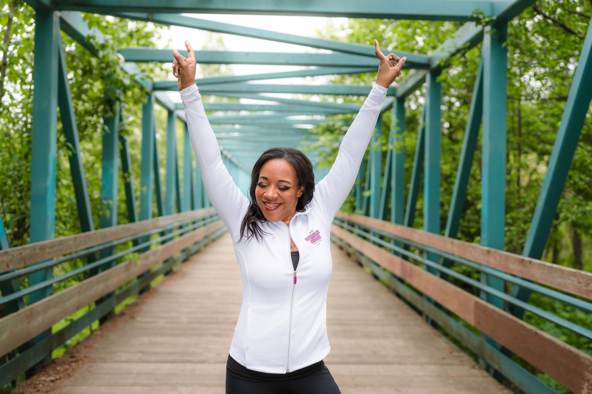 Woman in white jacket raises arms on a green bridge, smiling outdoors.
