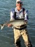 Man in fishing gear holding a silver fish in shallow water, smiling.