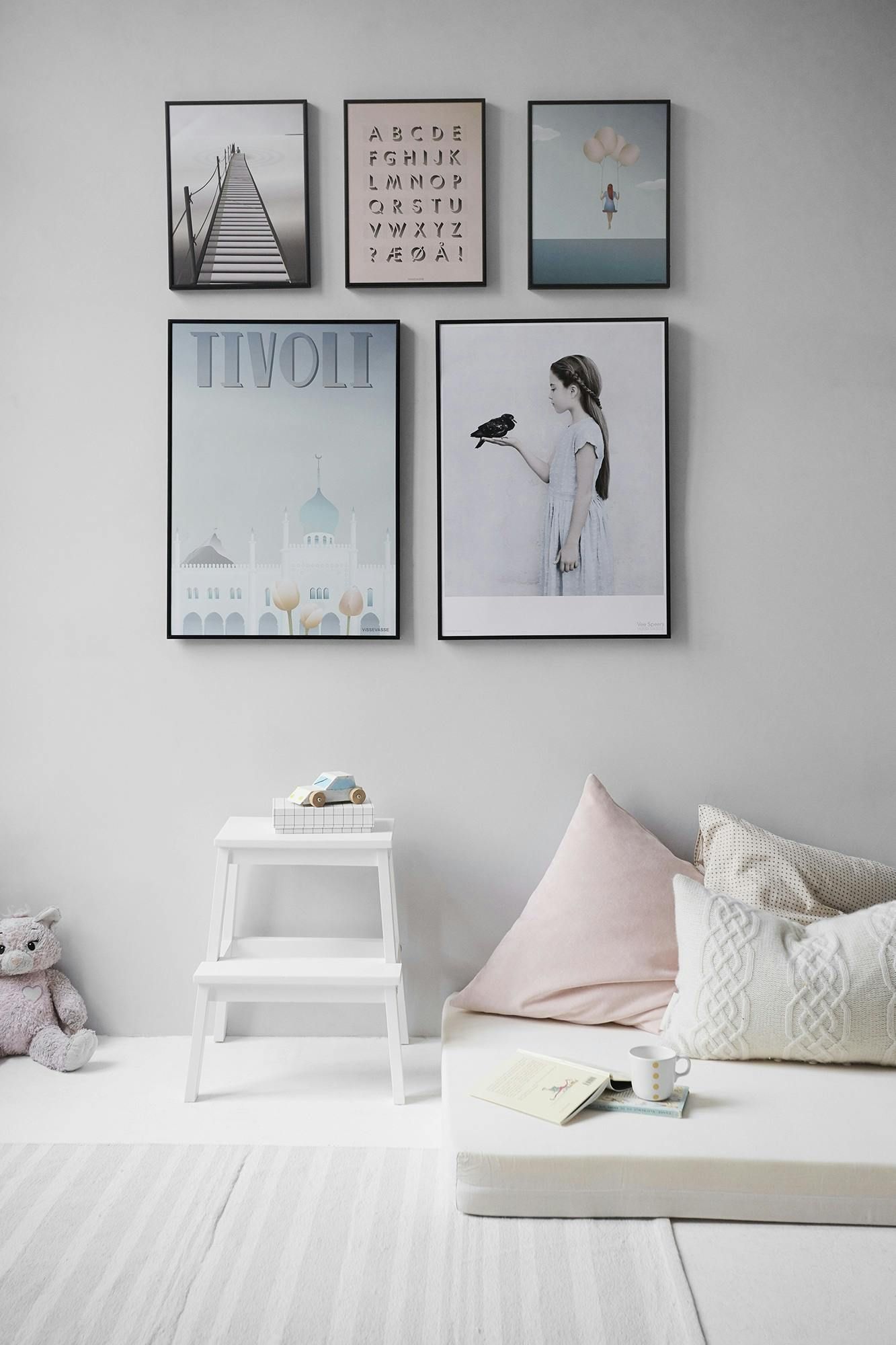 A child's room with framed art, a step stool, and floor cushions in shades of white, pink, and gray.