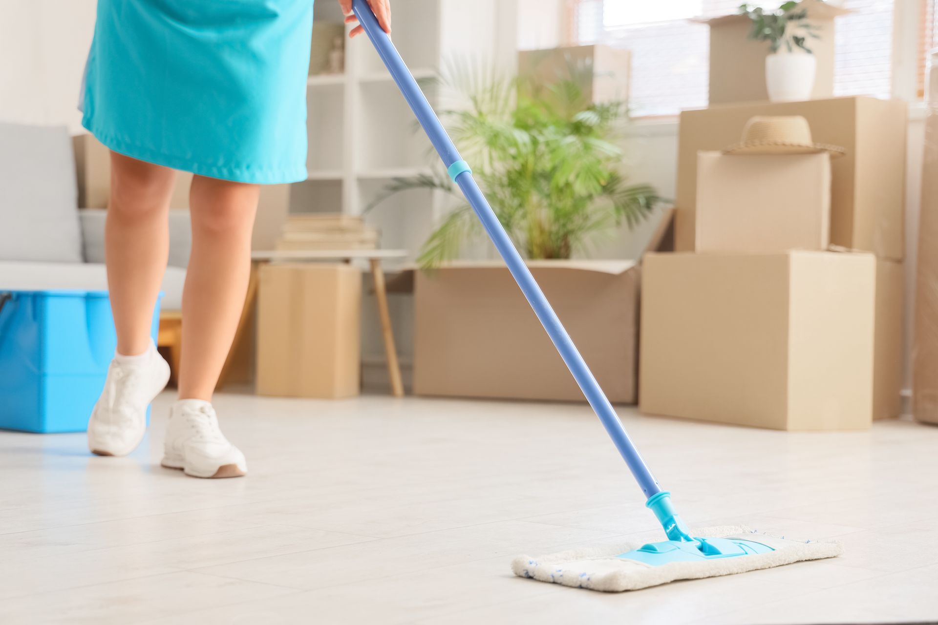 Woman mopping a light-colored floor, boxes in the background. She is wearing a blue dress and white shoes.