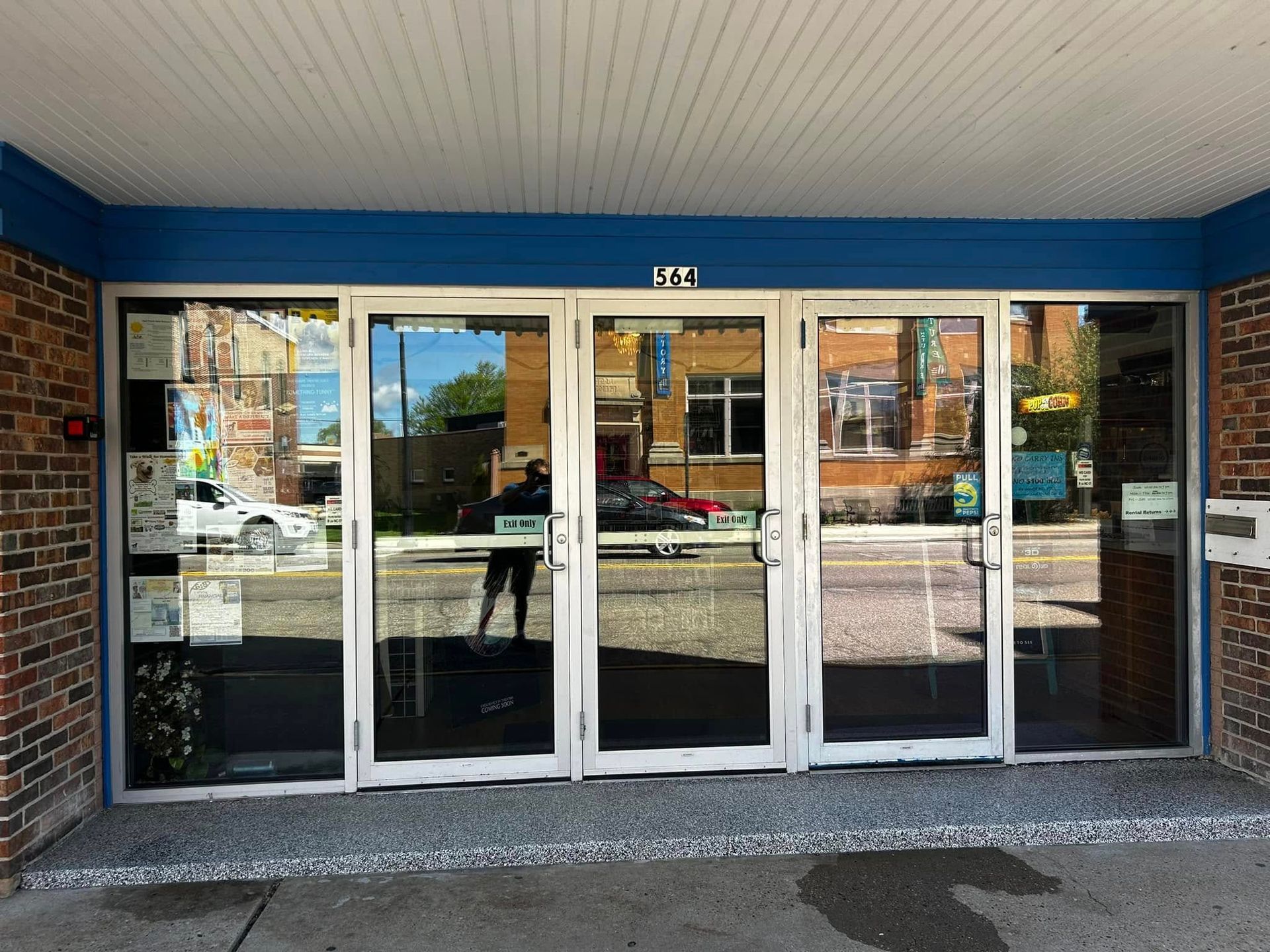 A storefront entrance featuring a set of four glass doors framed in blue under a white awning, with brick side walls.