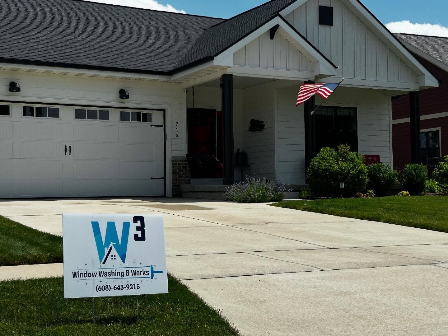 A white house with a black roof and a lawn sign for a company called W3, with a small American flag by the front porch.