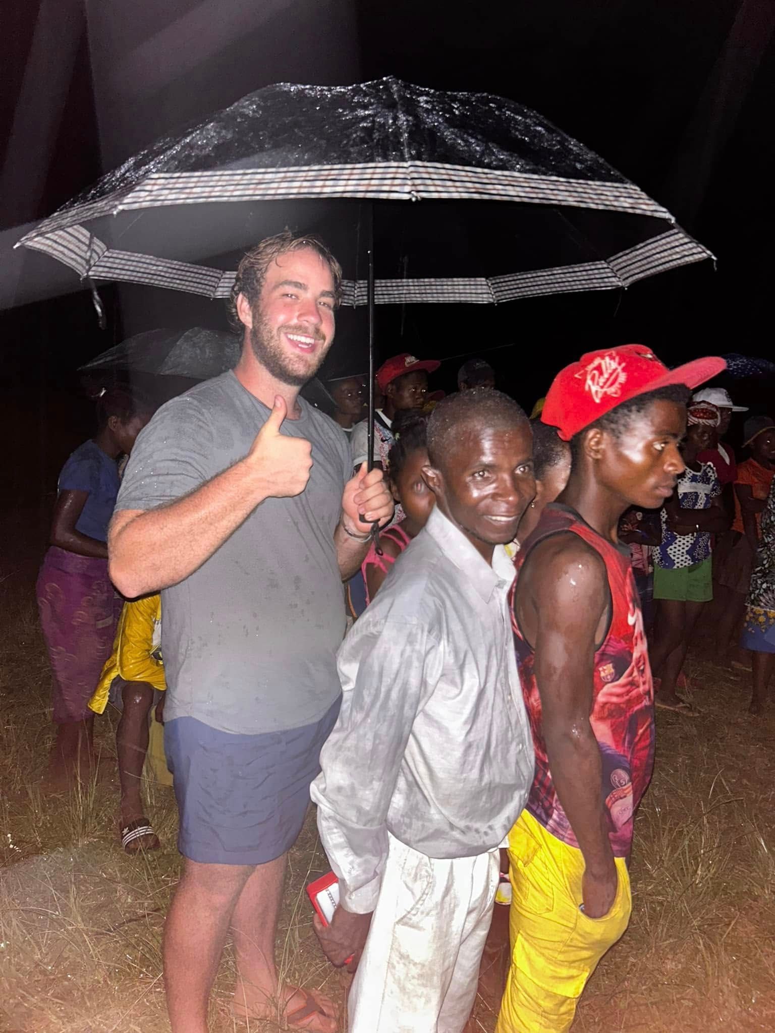 Man with thumbs-up under an umbrella, standing with a group of people in the rain.