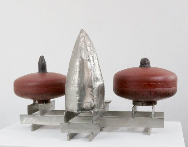 JOHN GIBBONS: ABSTRACTION AND BEING HUMAN
Visitors entering London’s National Portrait Gallery during the six months between mid-September 2009 and mid-May 2010 were confronted by five mysterious objects on the wall at the top of the long stair leading to the second floor galleries:  an installation entitled John Gibbons: Portraits.  The small, provocative exhibition was part of the NPG curator Paul Moorhouse’s “intervention series,” a program focusing on what are described as “twentieth-century artists who have developed innovatory approaches to portraiture” that has included such surprising works as a series of overscaled bronze heads by Anthony Caro (Bacon,Warhol).  As Moorhouse himself acknowledges, the Irish-born, London-based Gibbons, who since the mid-1980s has steadily declared his presence as one of Britain’s strongest and most original abstract sculptors, might seem an unlikely candidate for a show of portraiture defined in the usual sense.  At the National Portrait Gallery, in fact, his enigmatic constructions, poised on narrow, projecting shelf's (brackets), seemed initially to have little in common with the unequivocal images of specific individuals that the museum is known for.  Gibbons’s exhibited works – three large, rounded “cages” made of stainless steel and two smaller, dark, chunky “containers” – seemed to posit ideas unrelated to portraiture’s traditional considerations of recognizable traits and overt reference to appearances.   Instead, they appeared to explore purely sculptural notions of the relationship of interior to exterior, of volume to surface, and of openness to enclosure.  Their varied surfaces, some sleek, some dull, some bearing the traces of effort, bore witness to a dual allegiance to the industrial and the made;  this simultaneous dependence on the inherent qualities of metal and the transformative effect of the artist’s touch forced considerations of the importance of materiality and physicality to sculptural expression.  Cumulatively, the ideas and questions proposed by Gibbons’s compelling works seemed to belong entirely to the province of abstraction, in general, and, in particular, to the realm of the kind of abstract construction in steel that he has investigated for almost three decades.  
Yet the five apparently abstract sculptures placed above us against the wall also announced themselves as watchful but unyielding presences.  Far from reading as dispassionate or inanimate, they seemed to be eerily alert, acknowledging anyone who approached them, yet at the same time, they appeared to withdraw from the viewer’s gaze, like self-sufficient individuals who turned inward, lost in their own thoughts, challenging all comers to penetrate their secrets.  When, on becoming increasingly aware of this latent, elusive anthropomorphism, we sought confirmation (or denial) in the titles of the works, we discovered that the three cage-like stainless steel pieces, all made between 2008 and 2009, were identified, suggestively, as Jane/E/And Still,  Your Story/White Blackbird (Olmec), and Gráinne/Saying Hello (Electric Brain).  Each of the three was a roughly oval volume just under three feet high – larger than life size, but clearly with the proportions, more or less, of a head.  Each of the three had a distinct character, in part because of the idiosyncracies of its form, in part because of its component materials.  Jane/E, for example, built of flat straps and planes, was the most elongated and rounded.  A relatively smooth, enclosed upper section was caught in a ribbony, open-work structure of narrow, mostly vertical, apparently subtle slats of steel.  The elegant form, combined with the delicate hatchings and incisions that animated the surface of the straps, along with a few notes of carefully inlaid copper, seemed strangely feminine, with associations with antique caps and bonnets.  Your Story employed similarly scarified but more robust bands and carefully adjusted planes to conjure up a squarer, “jowlier,” more masculine form – as we should have expected from the reference to the massive stone heads of the pre-Columbian Olmec culture in the subtitle;  a short, centralized stack of copper plates made the  Your Story confrontational and a little truculent.   Gráinne, the visually lightest and most open of the three, seemed to have been momentarily formed out of strings of cylindrical stainless steel “beads” of varying dimensions that arc-ed, bent, twisted, zigzagged, and magically stopped, frozen as they described a loose, swelling form.  Even though we could penetrate the forms visually – seeing completely through them, in fact, was unavoidable – the sculptures retained a potent sense of exterior and interior.
This kind of paradoxical evocation of inside and outside, as well as mass and volume, by means of structures that are, in fact, completely transparent, first appeared as one of Gibbons’s major preoccupations during the late 1990s and the early 2000s.  He produced a powerful series of lattice-like works, carefully constructed with layered bars of metal – or, on occasion, of woven flat bands – which, despite being completely penetrable by the eye, had the unignorable presence of monoliths.  Often, the literal openness of their construction notwithstanding, we felt constrained to visually enter these uncanny constructions first through the larger, framed openings that Gibbons made in his grids, openings that somehow announced the difference between exterior and interior, underscored the notion that the volume described by the transparent exterior was, in fact, substantial and weighty, and made the distinction between surface and what was contained immensely significant..  Yet even as we acknowledged Gibbons’s cages as fictive masses, we became deeply involved in the nuances of the open grids that enclosed those illusory forms.  Variations in the thickness and the spacing of the bars, how things touched, what was in front of what, all assumed enormous importance.  Among the most impressive of these sculptures were paired of rectangular, boxy baskets woven out of flat straps of contrasting materials – stainless steel and copper.  Like the heads at the National Portrait Gallery, they seemed somehow pregnant with meaning despite their minimalist associations.  One of the largest, Om, 2000-02, balanced the “baskets” side by side on a wheeled platform, suggesting contingency and the possibility of change, as well as connecting us to the everyday world, through associations with utilitarian, industrial objects;  by contrast, a smaller, tabletop iteration of the theme, As One, 2001-02, evoked ritual objects or reliquaries, perhaps because of its intimate size.  Gibbons’s hand was visible in all of these sculptures, meticulously formed, as they were, by virtuoso technique and a certain amount of obsessiveness.  Looped ends of elegant, round bars, shifts in layering or in spacing, in works such as Light, 1999-2002, along with slight irregularities in the deployment of the flat metal bands in works such as Om and As One, forced us to consider the physical history of their construction, just as the seams and marks in Gibbons’s heads did.   But the more we concentrated on the material qualities of the exteriors of these constructions, the more convinced we became of the reality of their interiors – the chunks of space they delineated.  Like the emphatic edge drawing in Jules Olitski’s spray paintings of 1960s, which called attention to the limits of the pulsing expanse of color and asserted the difference between what was the painting and what was not, the bars and bands of Gibbons’s cages stressed the difference between the visible space within the lattice boundary and what was outside of it – that is to say, between what was the sculpture and what was not.  
Large sculptures of this type recalled both standing stones and human presence.  Just as the open volumes at the National Portrait Gallery clearly asserted themselves as heads, as well as abstractions, stalwart constructions like Rhythm/Play and Until, both 2000, conjured up the body.  Upright, confrontational, proportioned like massive torsos, they demanded that we considered them as personages as much as, or more, than objects.  Other works of this period, similarly proportioned and similar in scale, but horizontally oriented, such as The Crib, 1995-96, and To Be, 1996-97, provoked recollections of beds, biers, or ritual furnishings of some kind.  Occasionally, Gibbons underscored the implicit narrative of such works, most dramatically in The Crib, when he placed a rough hewn wooden stool beside an ambiguous hooded structure, unequivocally invoking human activity and habitation, and sounding a bracing “wrong” note of the anecdotal, certainly of the unexpected.  
Gibbons has said that he was wary of revealing such allusions for a long time, using them as part of his process, to animate the evolution of his sculptures, but resisting making them wholly visible.  Eventually, however, he felt compelled to allow them to become explicit.  Once he “opened the door,” as he put it, he found the enriched layers of meaning that attached themselves to his sculptures to be, paradoxically, liberating,   It was as if acknowledging the multivalent sources of the things that he made gave him permission to tap into still more deeply felt experience, both emotional and perceptual.  By the mid-2000s, he intensified his exploration of ways of alluding to the corporeal and the physical in sculptures that were as opaque and seemingly impenetrable as the caged and woven works were transparent:  irregular masses, apparently created by wrapping and folding intractable sheets of stainless steel.  (The sense of “shrouding” and “protecting” inherent in these works, as well as their intricacy, may have had something to do with the fact that the stainless had originally belonged to David Smith, a sculptor Gibbons reveres and whose “delicate” touch he has often praised.)  
In the stainless “packages,” Gibbons’s masterly ability to manipulate his materials came into play, yet we were engaged by the delicacy with which the famously recalcitrant metal was scored, bent, or pieced together only after we had begun to ponder the suggestion of something invisible and unidentifiable contained within.  In small works of this type, such as A Thousand Dreams, 2005-07, or She Said/…., 2007-08, it is impossible to guess at the contents of the enigmatic “parcel;”  the addition of colored vinyl “strapping” on some of these sculptures intensified the sense of containment and added an unexpected element of both literalness and discord, like the stool in The Crib.  Larger works of this period were both more forthcoming and more explicit.  Tall, vertical, opaque sculptures such as Presence, 2005-07, with its form-enhancing belt, insisted on being read simultaneously as vaguely architectural and as slim, armless torsos.  Their proportions and orientation made them body-like, absent any literal representation of the body, in same way that the portrait heads were about specific individuals without referring to their appearance.  Like the transparent works that preceded them and the allusive “portraits” that followed them, Gibbons’s opaque, impenetrable sculptures also forced us to think about the difference between exterior and interior – about volume as something defined and contained by unpredictable structure.
The installation at the National Portrait Gallery made clear that this kind of oblique reference and insistence on the metaphorical possibilities of interior space has long been part of Gibbons’s practice, even when he seemed fully engaged by abstraction.   Mounted below the large, open heads were two small, angular sculptures made of dark, patinated steel, dated 1981-1984, unequivocally labeled with the names of Gibbons’s children, Portrait of Sharon and Darragh’s Place.  Rather than suggesting heads or bodies, they insistently recalled reliquaries and tabernacles, or miniaturized buildings, with all abrupt transitions and sharp junctures softened into subtle transitions.  At the time, Gibbons was making large, enclosed sculptures like confessionals or shrines, scaled to the body, impossible to enter and impenetrable to the eye but redolent of secret, inhabited places within;  their vaguely architectural vocabulary could be read as alluding to the church furnishings of his Catholic upbringing in the West of Ireland.  As Gibbons explains it, in making Darragh’s Place, he didn’t intend from the start to make a sculpture about his son, but rather recognized qualities in the evolving work that made him think about it as a head of the boy – “a container,” as Gibbons puts it, “for his personality.” “I’ve been thinking about the head as a volume, as a container for ideas and thoughts, a container for experience,” Gibbons told me recently.
The titles of his recent abstract volumes at the National Portrait Gallery turn out to be quite literal;   each sculpture was conceived – or at least recognized – as embodying a particular person, evoking a particular personality.
Yet it would be disingenuous to deny that, certainly at first acquaintance, the formal differences between the small “reliquary” portraits of Gibbons’s children on view a the National Portrait Gallery and the generously proportioned portrait heads seemed greater than their similarities of intent.  At the same time, tracing the evolution of Gibbons’s work makes it clear that the acknowledgement of human presence, whether by allusion, metaphor, or implication, turns out to provide a continuous thread, from the “confessional,” “shrine,” or “reliquary” sculptures of 1980s to the cage-like heads of recent years.  The proportions of the human body inform of almost all of Gibbons’s efforts, obliging us to consciously or unconsciously measure his sculptures against our awareness of ourselves.  This animating sense of human dominance manifests itself in even the most architectural and seemingly non-referential of Gibbons’s sculptures, not only the “confessionals” of the 1980 but also a series of generous, extended constructions distilled from industrial structures – walkways, railings, entrances, walls, and the like – that devolved from them in the early 1990s and triggered the cages.  All of these works resonate with memories of our gestures, the length of a stride, the reach of an arm, so it comes as no surprise that Gibbons has says that his work, however apparently abstract or concerned with the physical and formal aspects of making three-dimensional objects, has always been “a reflection on the nature of being human.” 
The works at the National Portrait Gallery made this more explicit than anything Gibbons has done in the past and make us eagerly await what he will do in the future.
Karen Wilkin
New York, March 2011 
