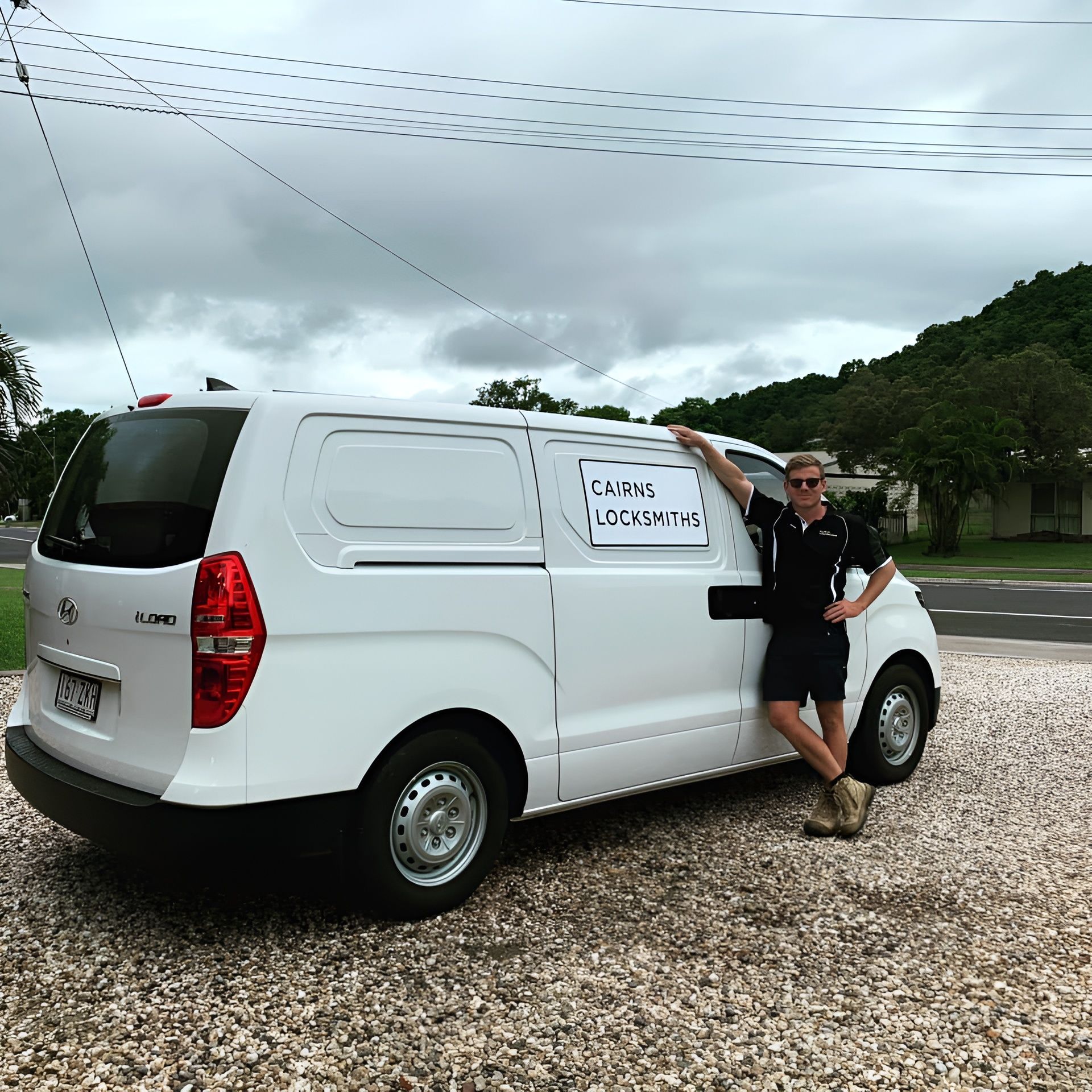 Man standing next to a white van with text. Cloudy sky, gravel ground, and greenery in the background.