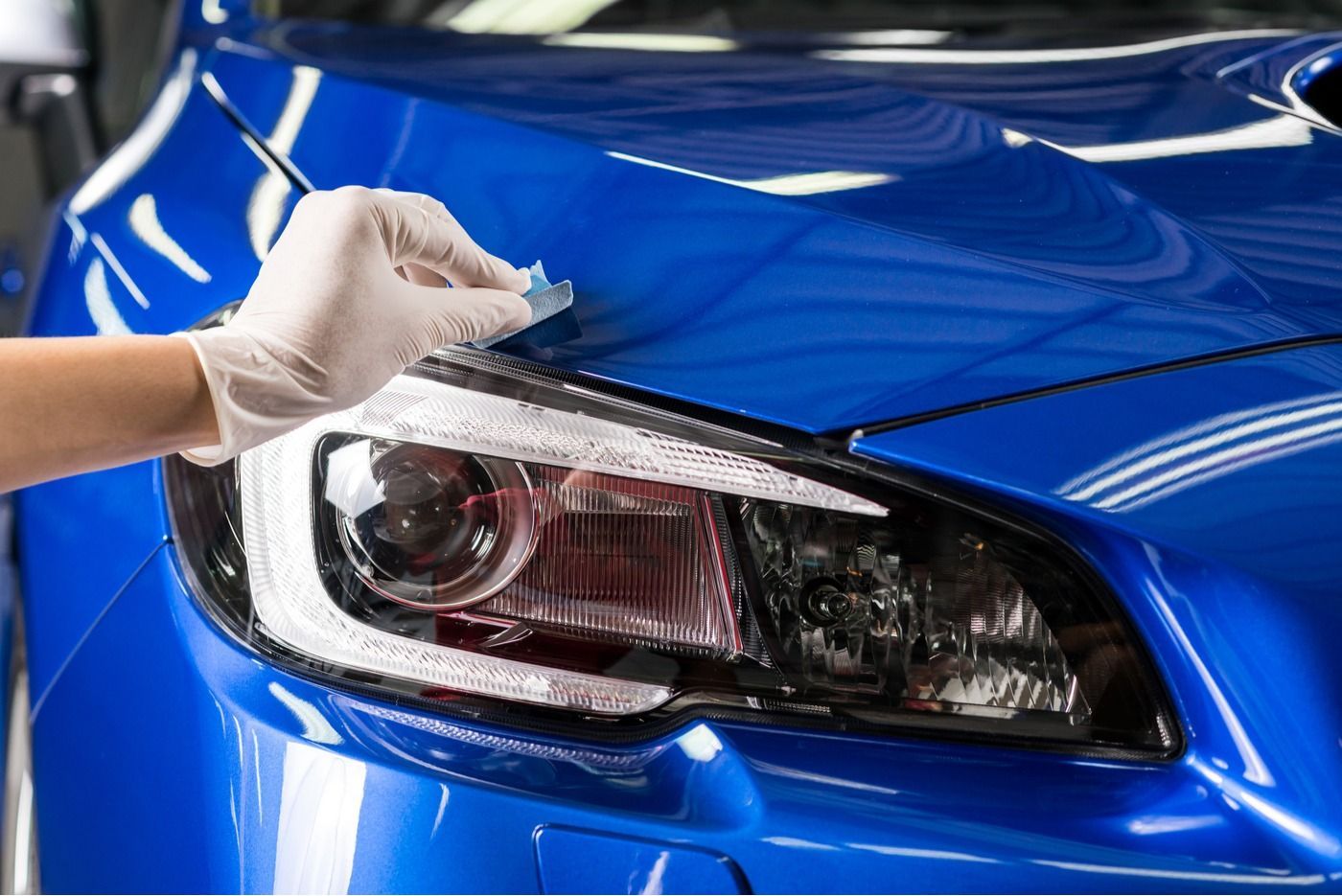 Gloved hand applying a protective film to a blue car's hood, near the headlight.