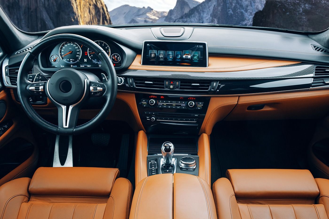 Interior of a brown leather luxury car, dashboard with display, steering wheel, and mountain view in the background.