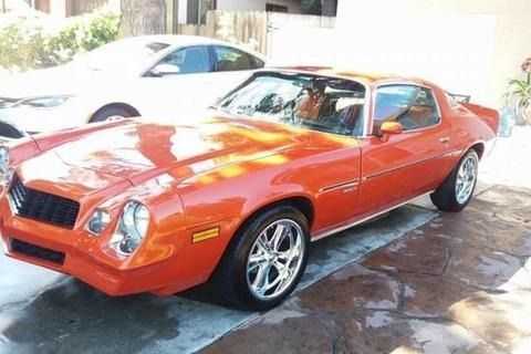 Orange classic car parked on a stone surface, shiny chrome wheels, sunny outdoors.