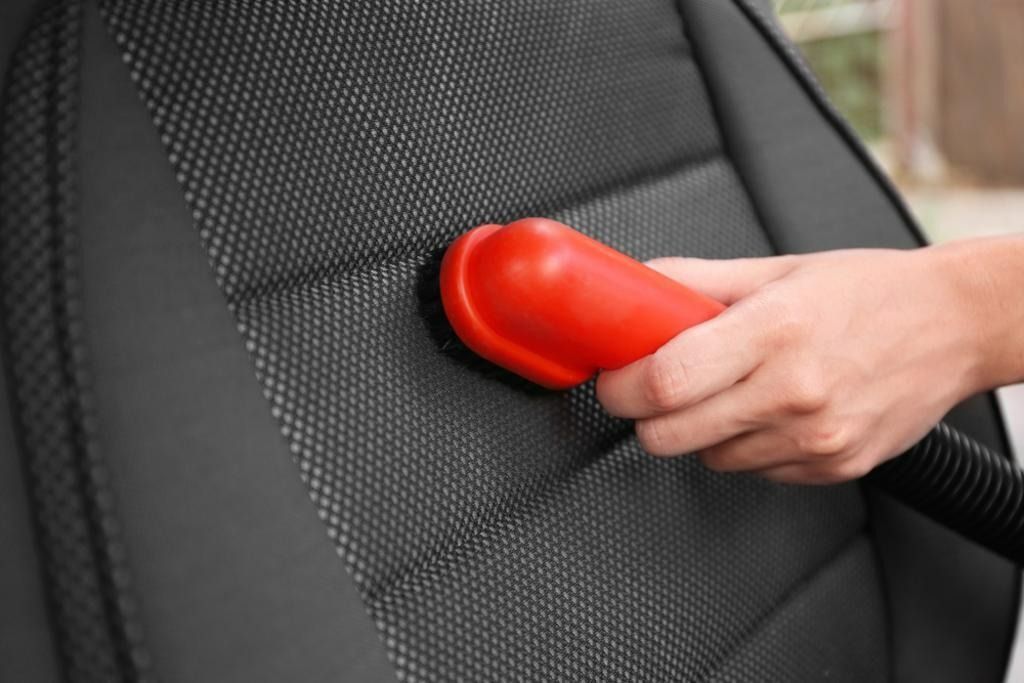 Hand vacuuming a black car seat with a red nozzle.