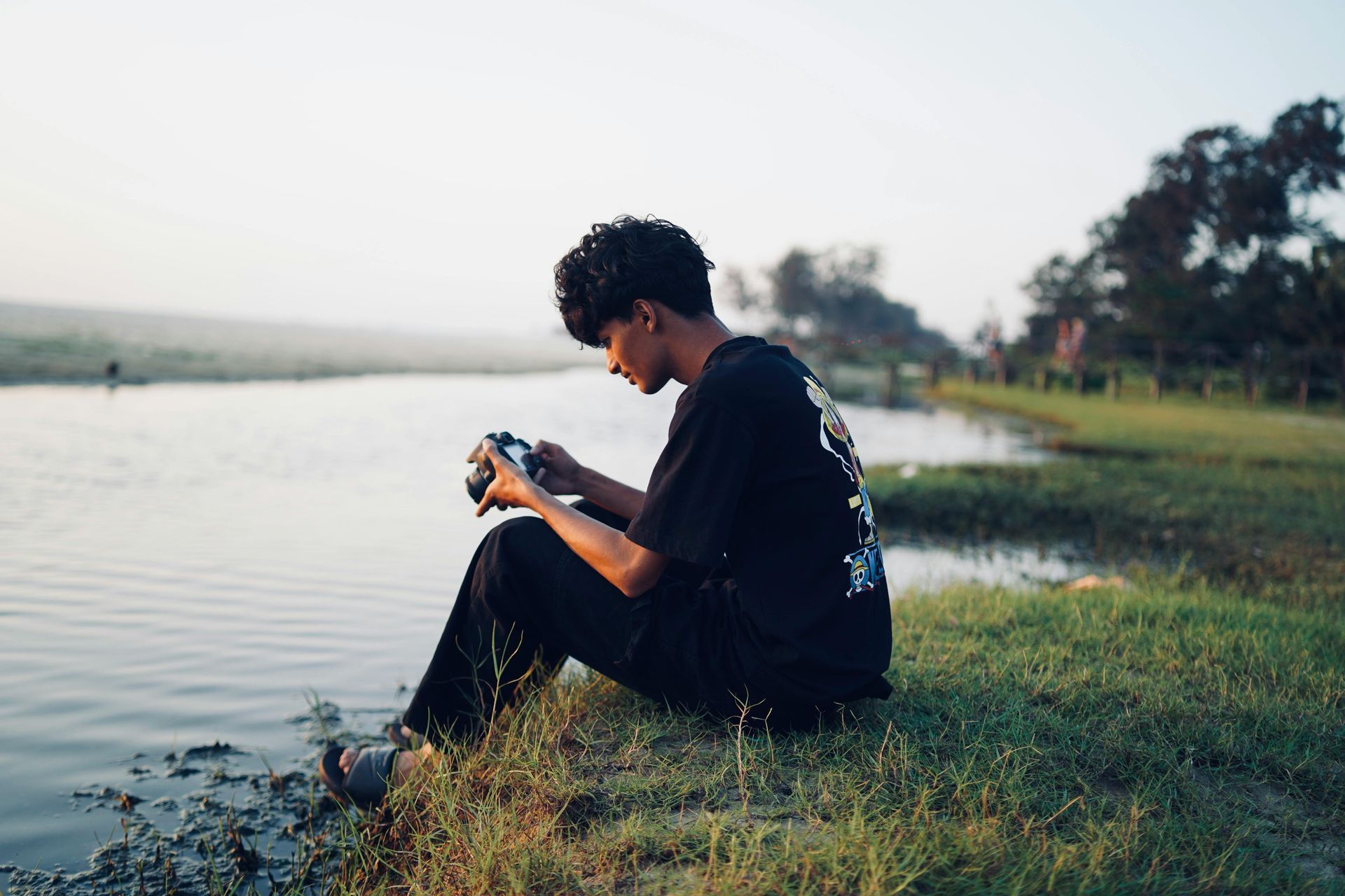 Een jongeman zit op het gras bij een meer en maakt foto's met een camera.