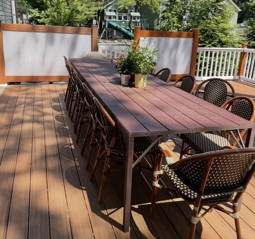 Long wooden table on a deck with rattan chairs. Plants in pots are on the table.
