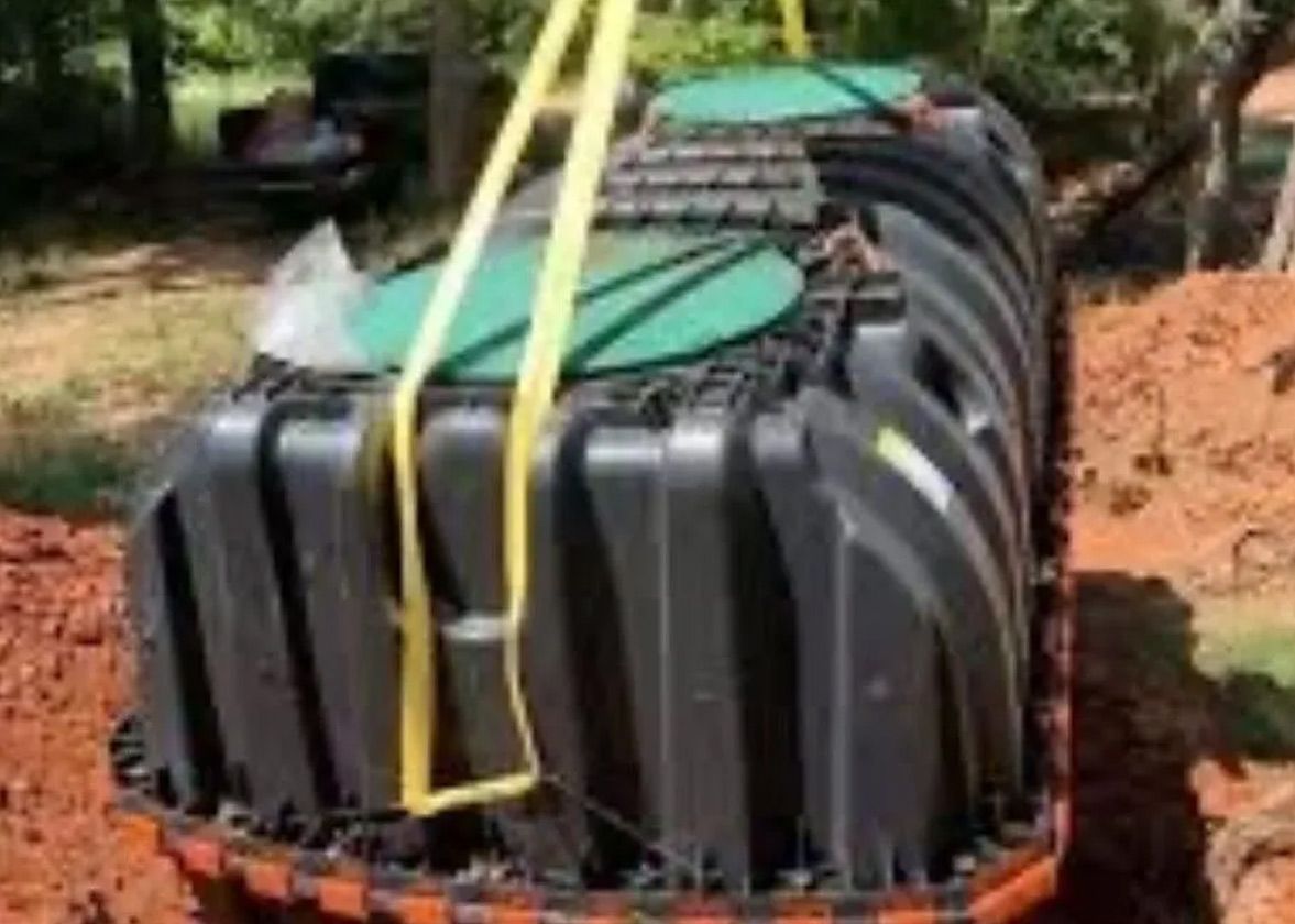Black septic tank being lowered into a trench, secured by yellow straps. Green lids visible.