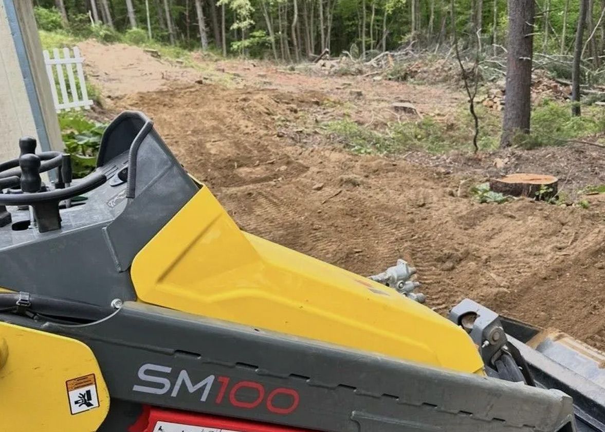 Yellow and gray skid steer (SM100) leveling dirt in a yard near a white fence and woods.